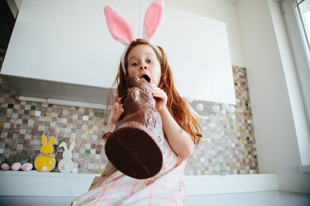 Red head girl wearing dress and bunny ears, sitting at the kitchen counter, holding chocolate bunny and expressing astonishment