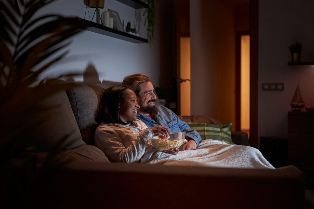 Side view of cheerful mixed race young couple at home watching movies at night in the sofa with a blanket and eating popcorns.