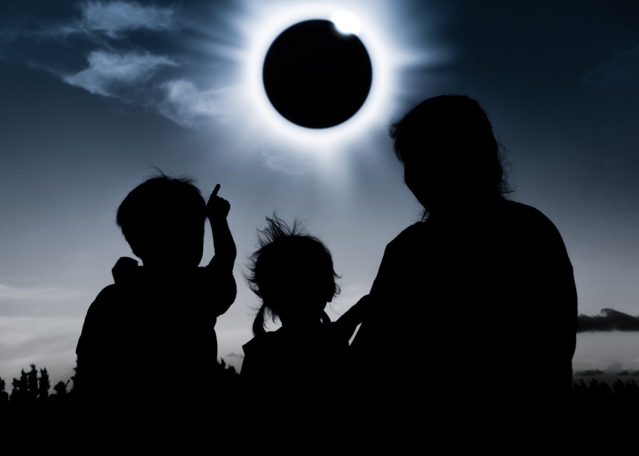 Natural phenomenon. Silhouette back view of mother and child sitting and relaxing together. Boy pointing to solar eclipse on dark sky background. Happy family spending time together. Outdoor.