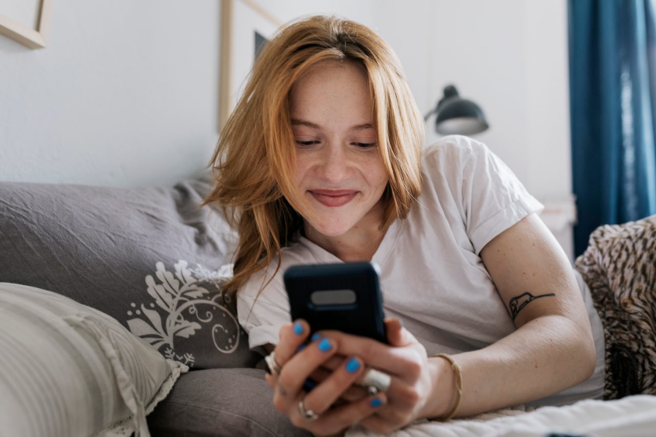 A woman smiling while lying on her bed at home and messaging on an online dating app using her smartphone.