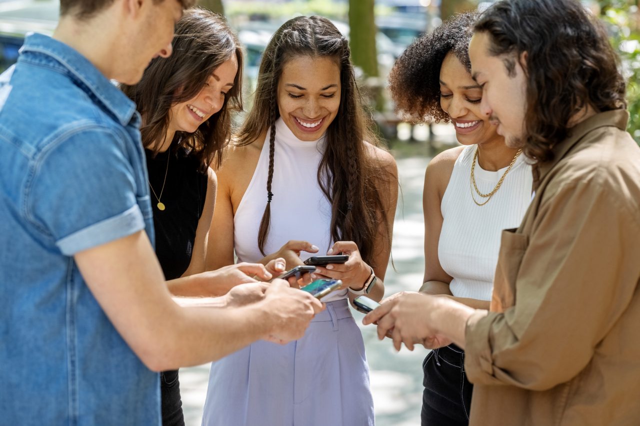 Group of young people looking at their smart phones outdoors in the city and smiling. Happy group of young friends having fun together in city.