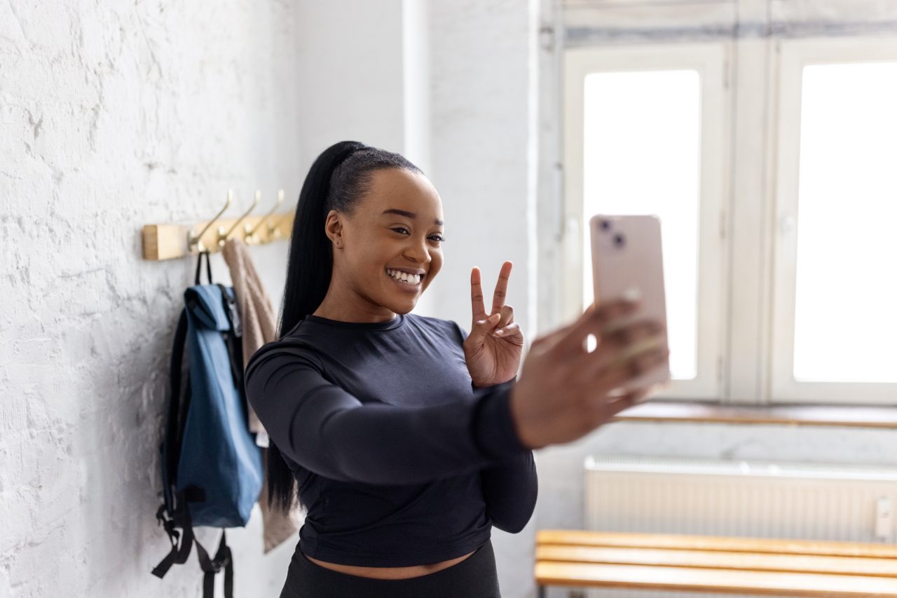 Young African woman taking selfie in the dressing room at gym. Smiling female using her cell phone for taking self-portrait in the gym locker room.