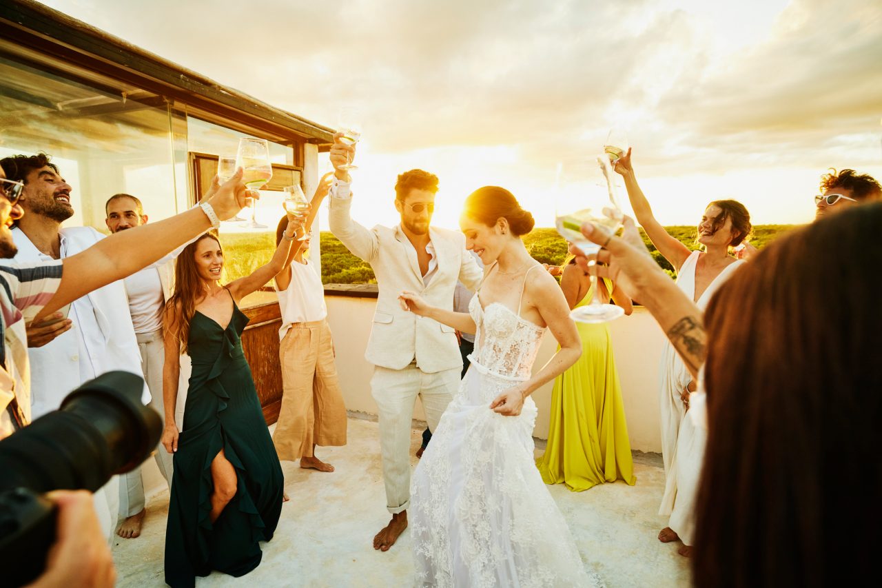 Wide shot of smiling bride and groom dancing and celebrating with friends on rooftop deck at sunset after wedding at tropical resort