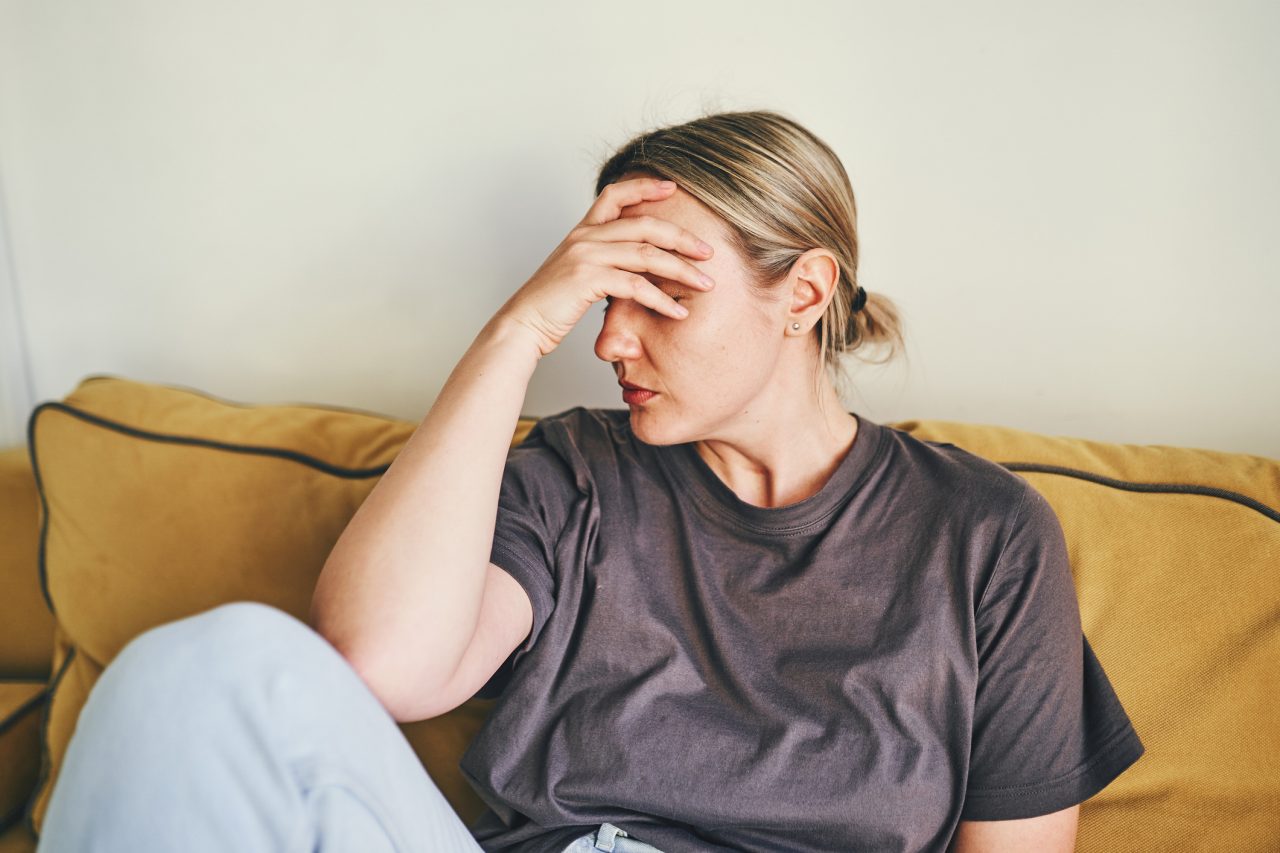 A young woman is sitting at home on a yellow sofa with her head in her hands, sad or showing a headache or frustration and depression. The concept of health problems , headache or stress