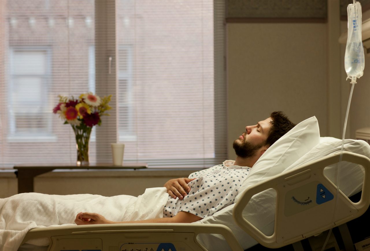 Bearded 20s man lying in hospital bed with eyes closed in pain.
