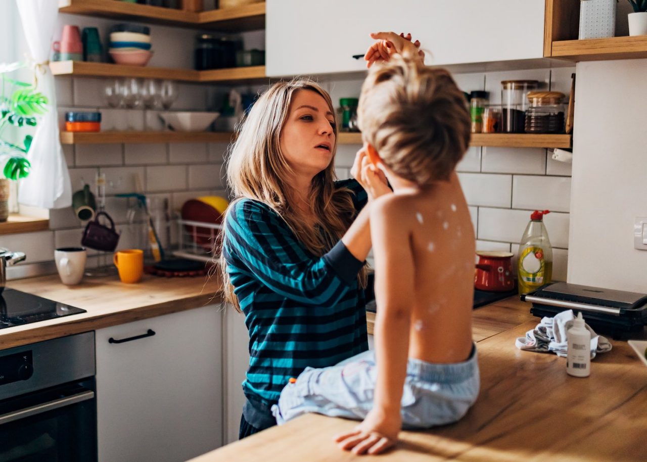 A  four-year-old boy sits on the kitchen counter with his arm raised while his mother applies measles medicine to him