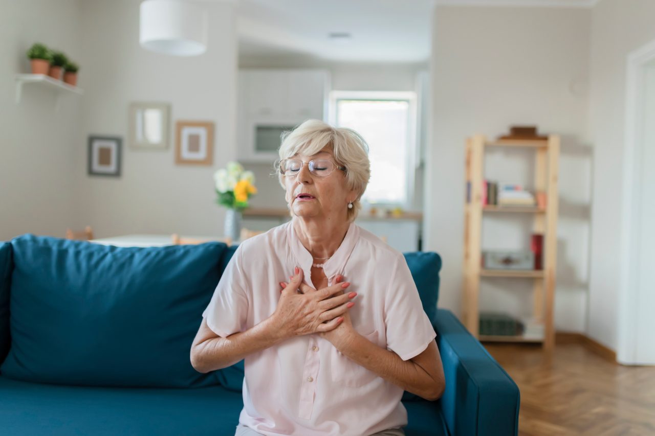 Senior Woman Suffering From Chest Pain While Sitting on Sofa at Home. Portrait of Elderly Woman Having Heart Attack